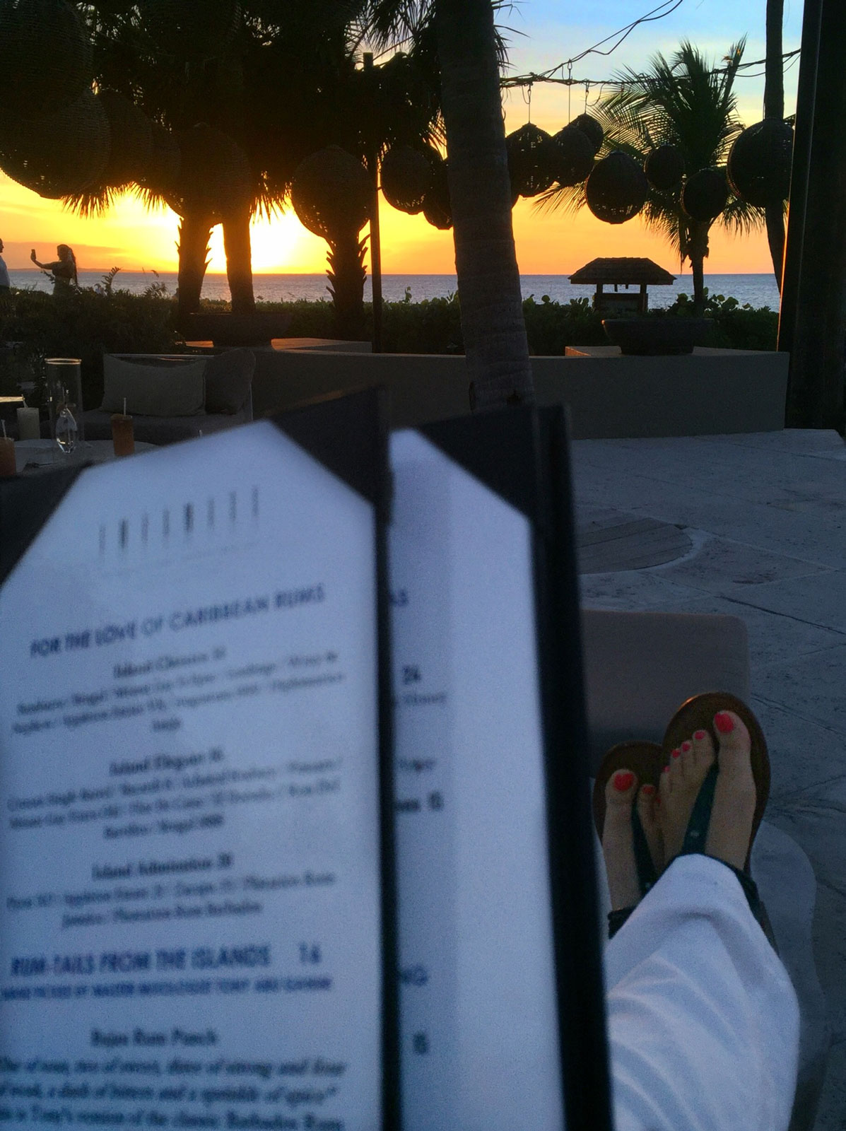 Orangish yellow sunset over the water with palm trees in the side and background. Taryn pinkish orange toenails are vibrant.