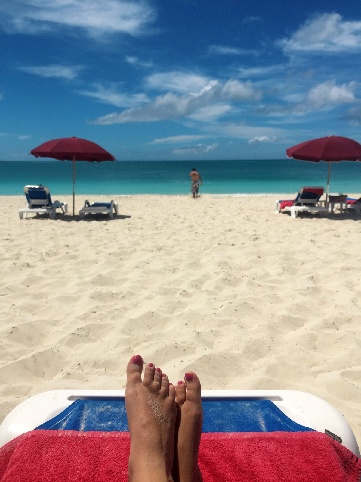 Taryn's view of the beach from her chair, you can see her feet and a lot of beach, then Brandon walking into the beach, lots of depth in the picture, a set of beach chairs with an umbrella on both sides of where Brandon is walking.